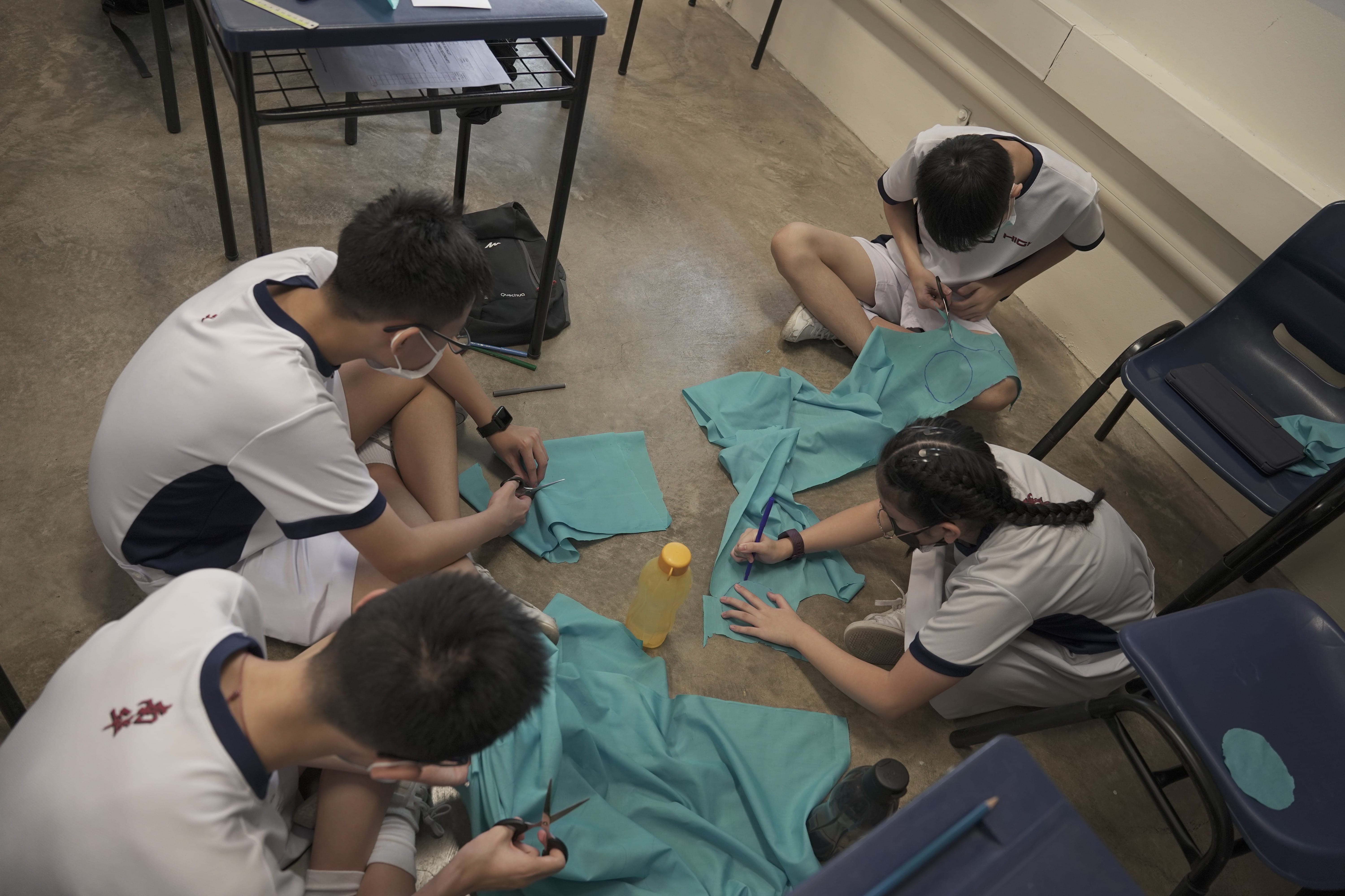 Four students in white uniforms sit on a classroom floor cutting and marking teal fabric with scissors and pens.